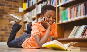 Afro-American kid leaning on the floor smiling with reading a book in the library