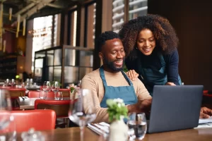 black men sitting in a restaurant table with a woman standing behind both looking at a computer doing business
